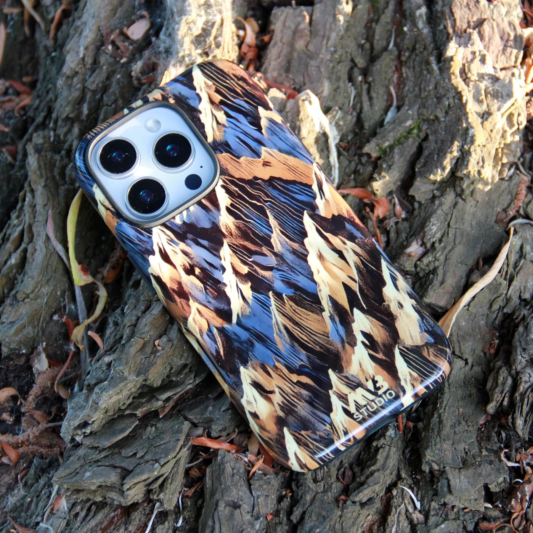 Overhead view of an iPhone 16 Pro Max featuring a distinctive blue and earthy-toned feather pattern case, placed on natural tree trunk texture. | MVB STUDIO STORE
