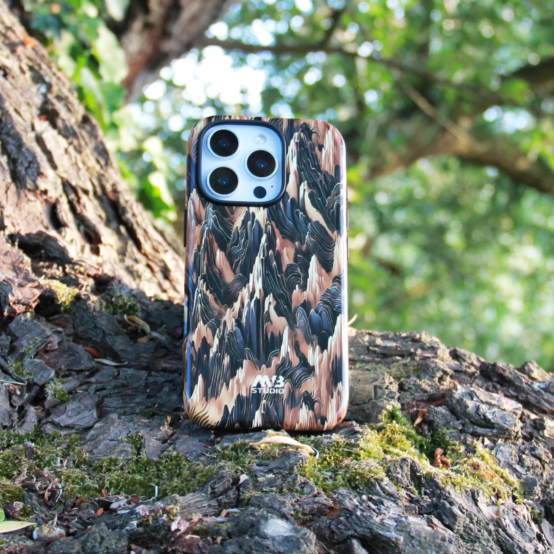 Close-up of an iPhone 15 Pro Max with a textured camouflage-style phone case standing upright on a mossy tree trunk in a forest setting | MVB STUDIO STORE