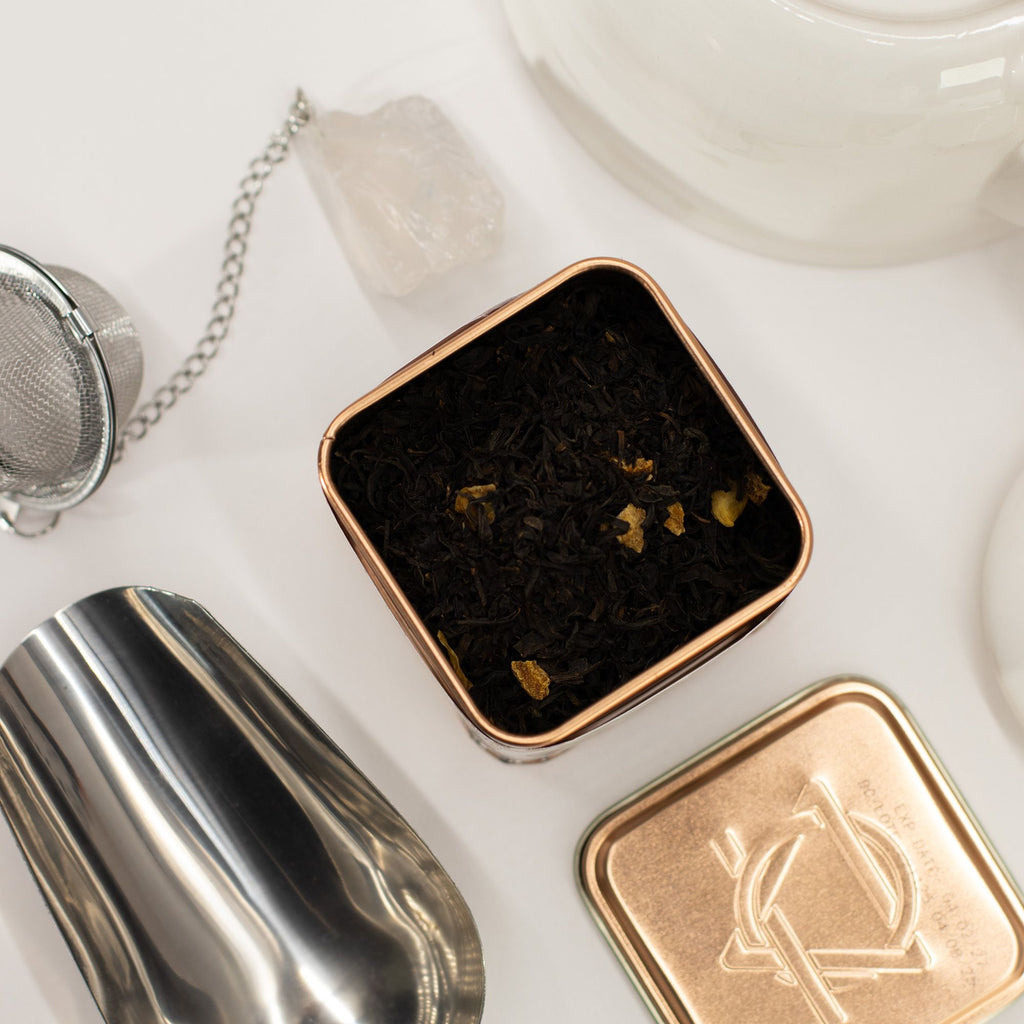 Black Tea with Orange Peel Citrus Loose Leaf Tea in a small square container with a silver tea strainer and white teacup in the background.