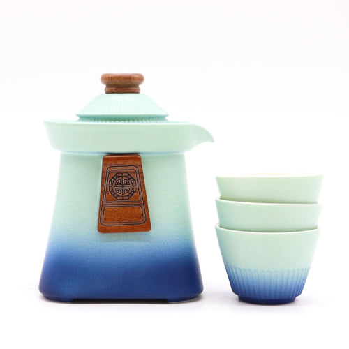 Gradient blue and green ceramic teapot with wooden lid and leather strap, accompanied by matching cups on a white background.