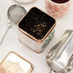 Tea tin with tea leaves Chai Black Tea, tea strainer, and cup on a white surface