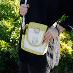 Person holding a yellow and white crossbody bag with greenery in the background
