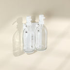 Two clear glass bottles with white spray nozzles attached to a beige wall.