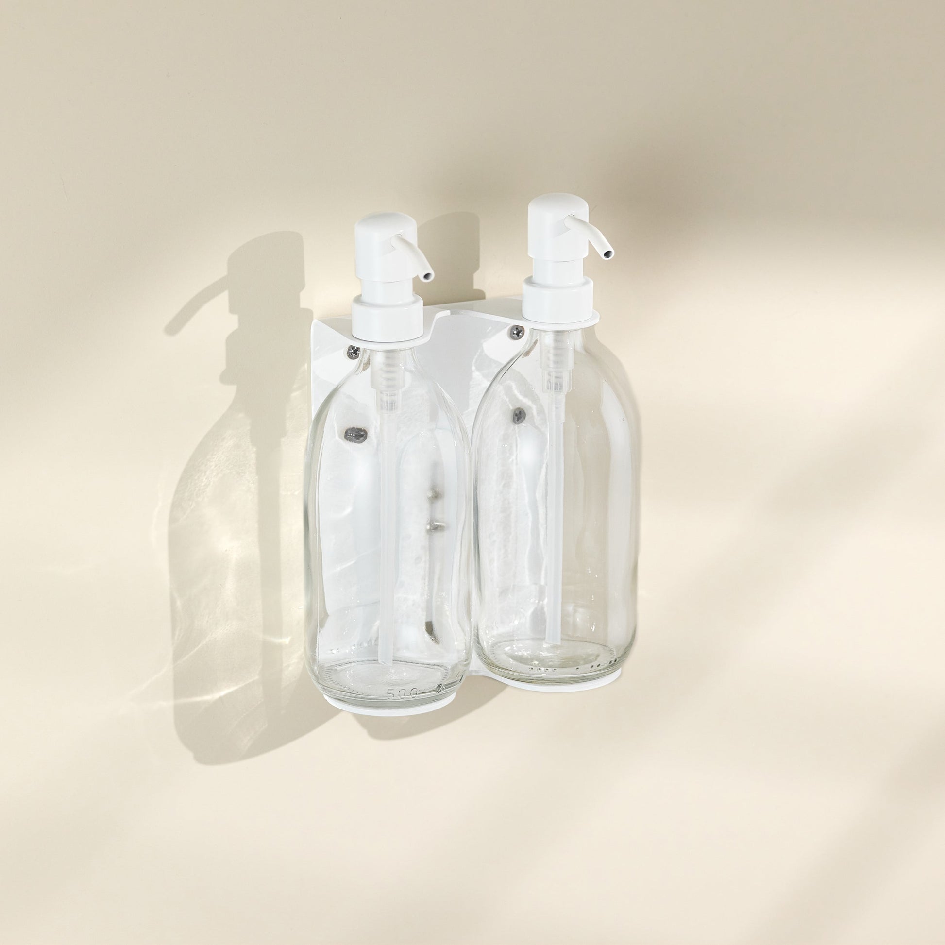 Two clear glass bottles with white spray nozzles attached to a beige wall.