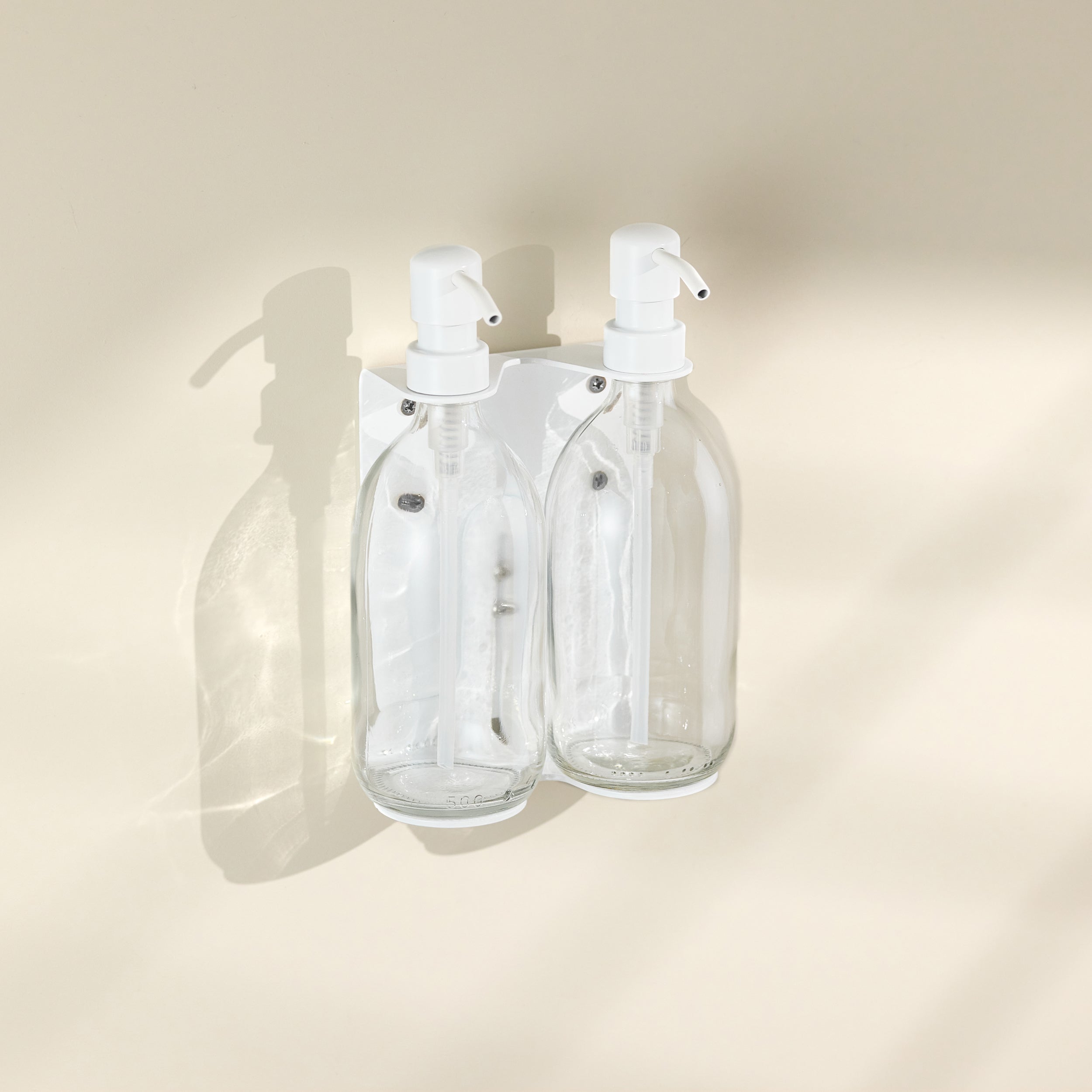 Two clear glass bottles with white spray nozzles attached to a beige wall.