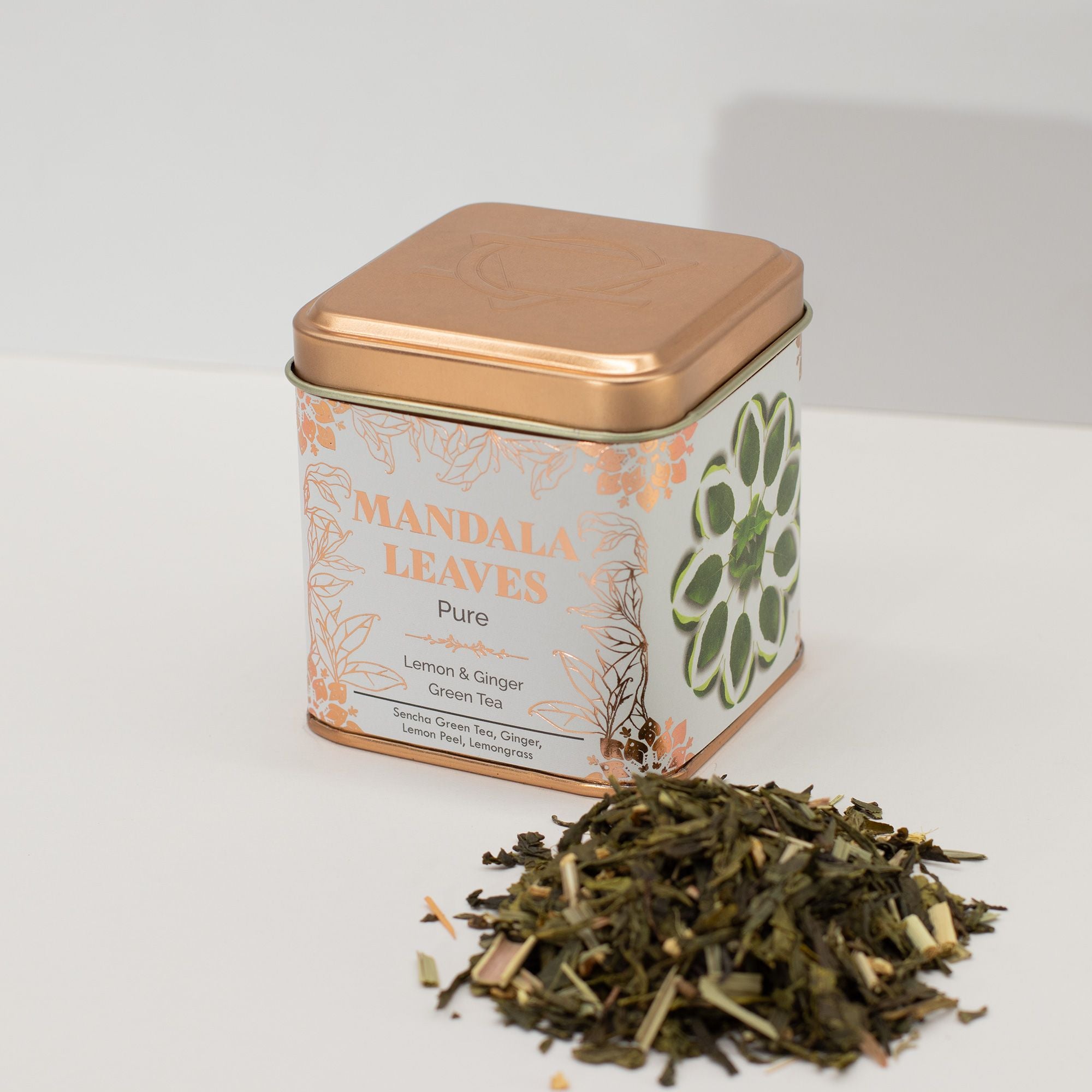 Tea tin labeled 'Mandala Leaves' with Ginger & Lemon Green Tea Sencha spilling out on a white background