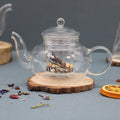 Clear glass teapot with dried herbs on a wooden coaster against a gray background
