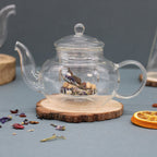 Clear glass teapot with dried herbs on a wooden coaster against a gray background