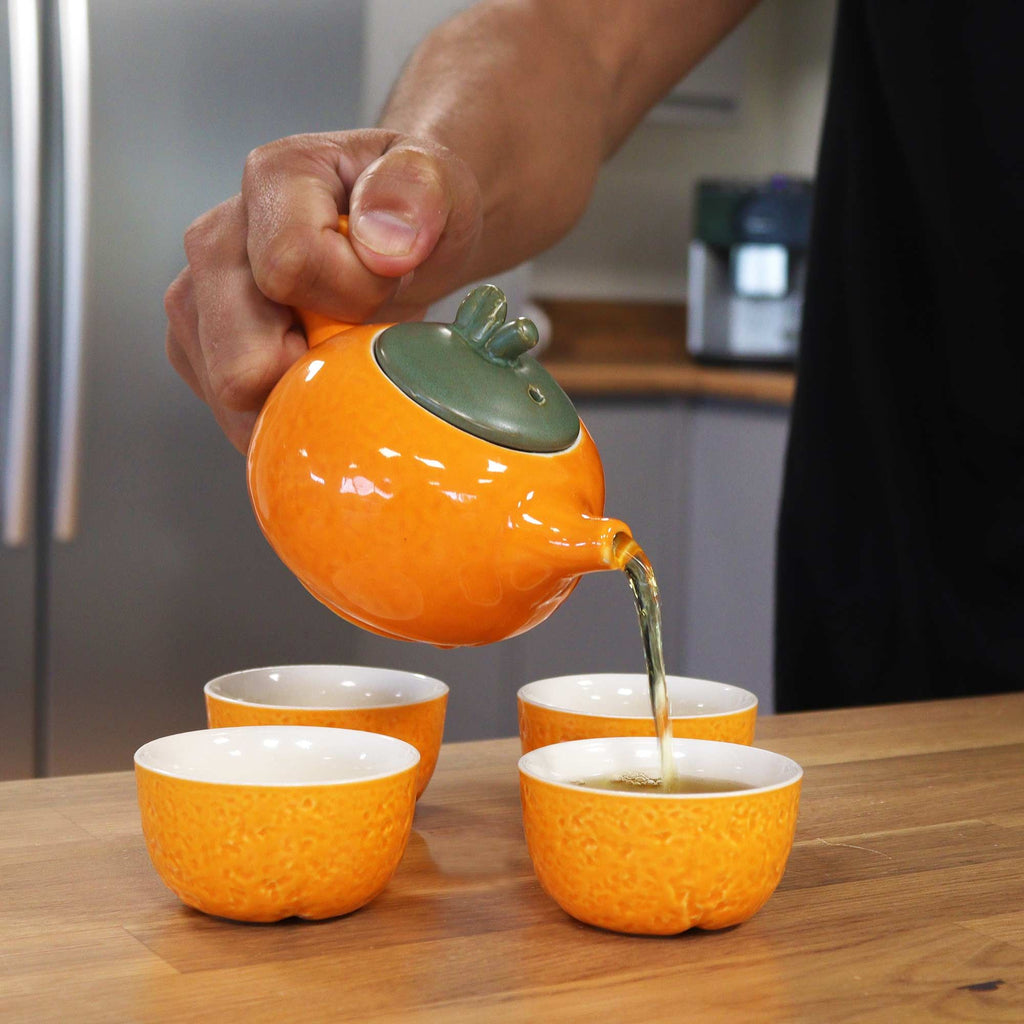 Person pouring tea from an orange teapot into four matching cups on a wooden table.