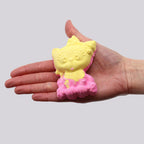 Hand holding a yellow and pink cat-shaped bath bomb against a plain background