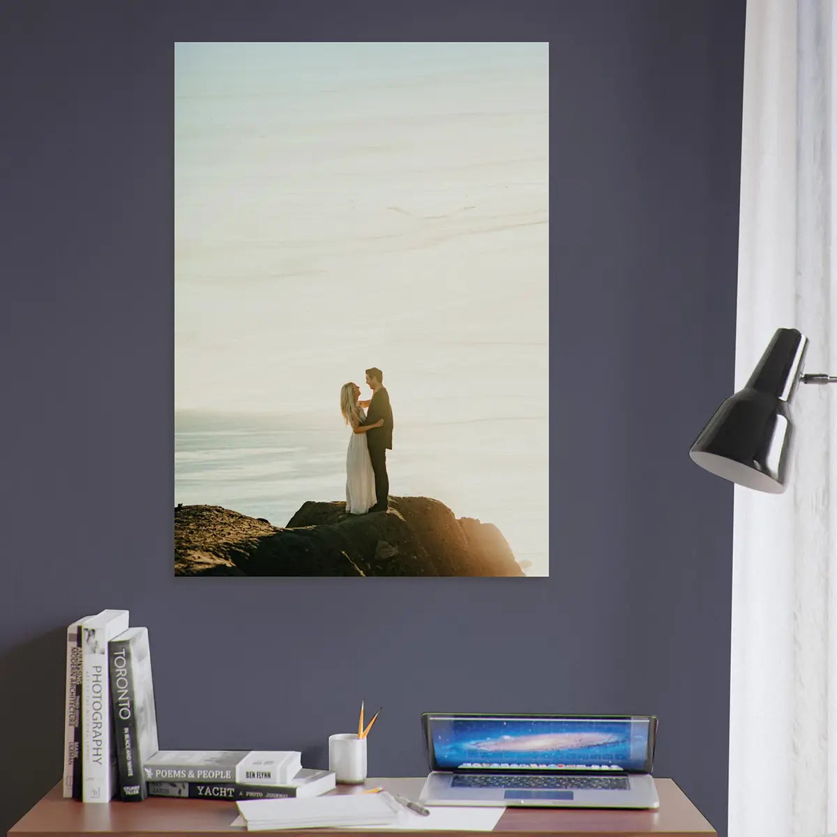 Wall art of a couple on rocks with a laptop and books on a desk below wood print.
