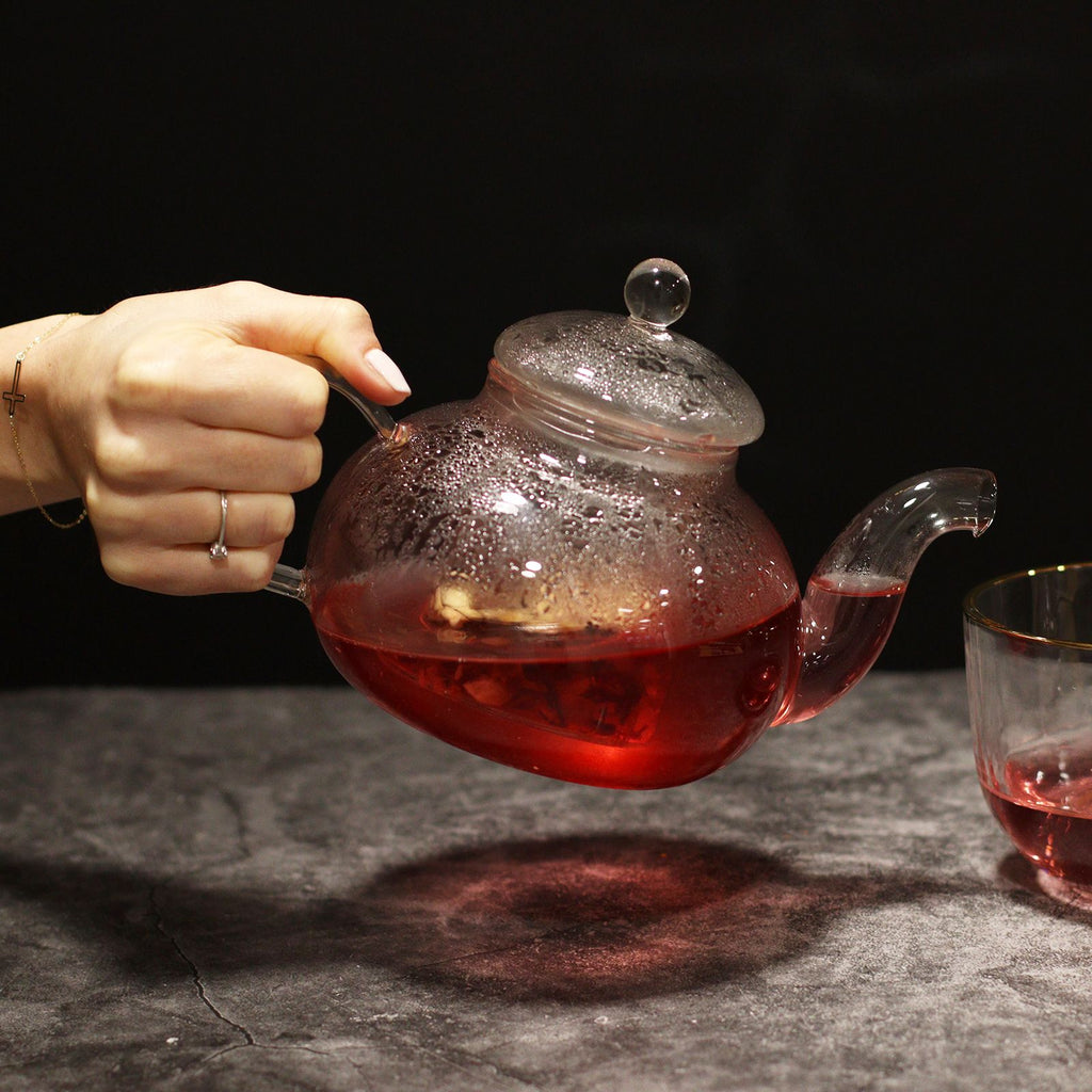 Hand pouring red liquid from a glass teapot into a glass on a dark surface.
