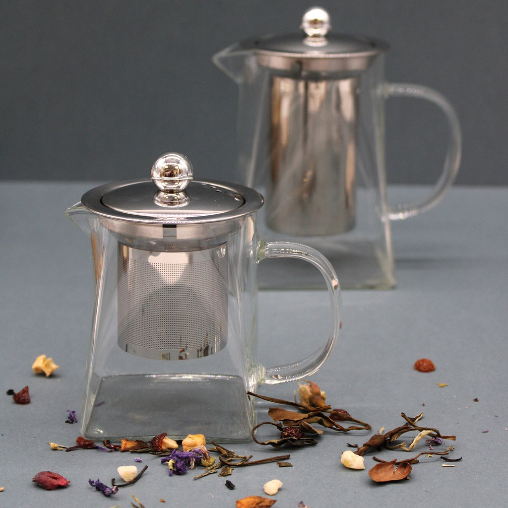 Clear glass teapot with metal mesh infuser and handle on a gray surface with dried herbs.