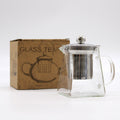Clear glass teapot with metal lid and infuser on a white background, next to a cardboard box labeled 'Glass Tea'.
