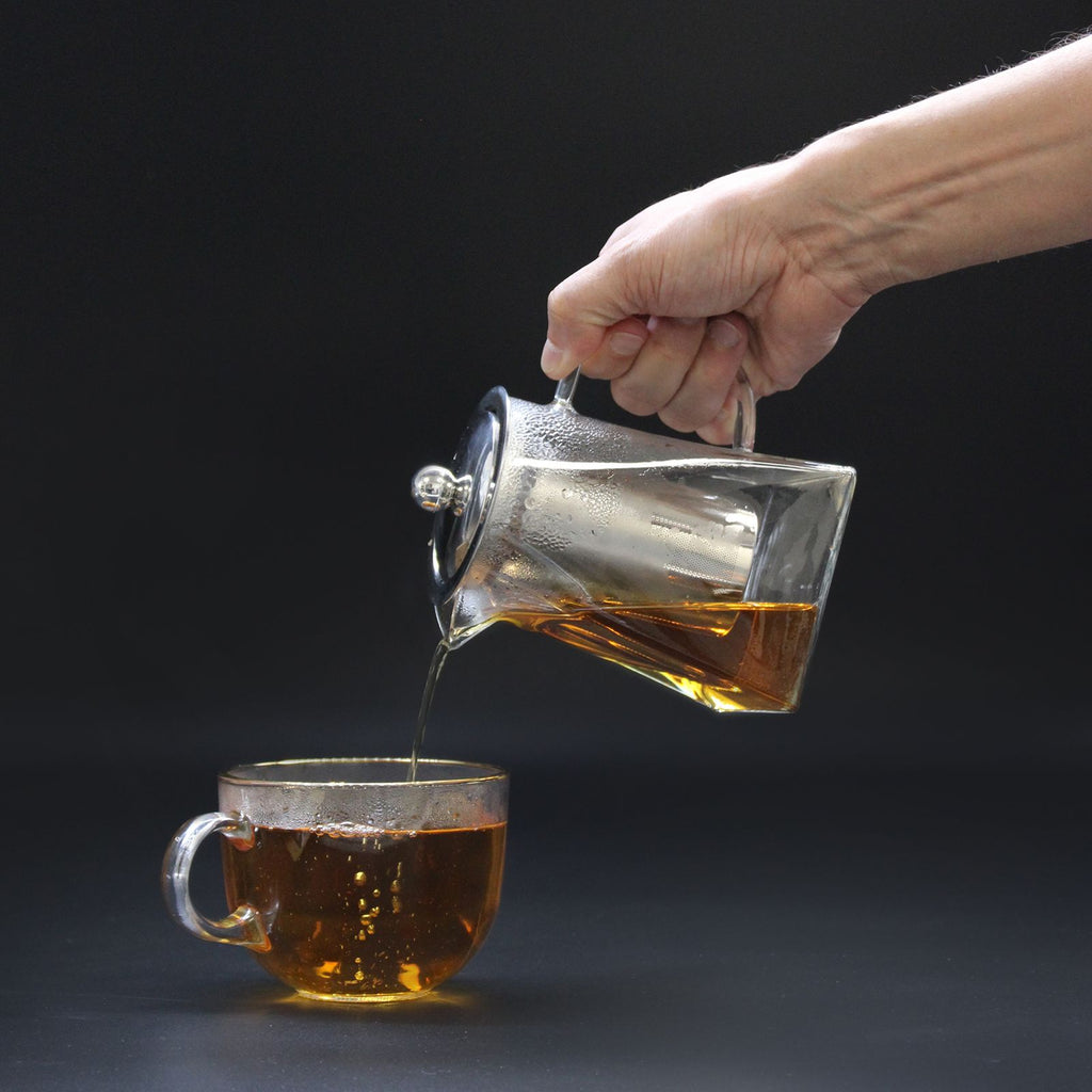 Hand pouring tea from a glass teapot into a glass cup on a dark background