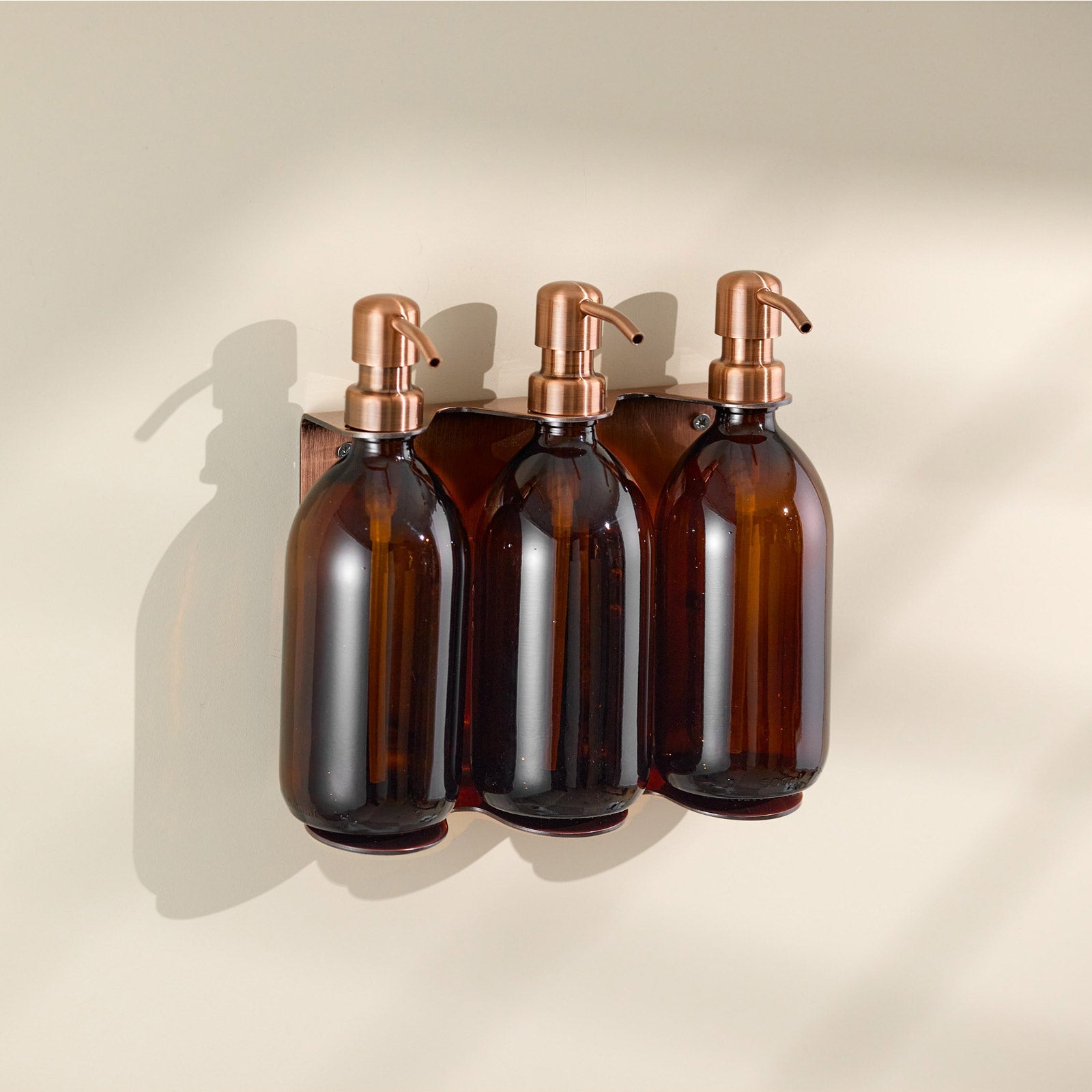 Three brown glass bottles with copper pumps on a beige wall-mounted holder.
