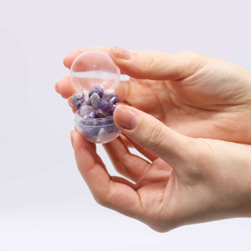 Hand holding a small transparent container filled with purple and gray stones against a light background