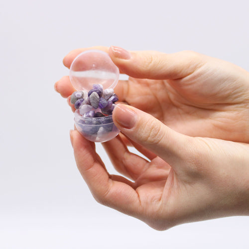 Hand holding a small transparent container filled with purple and gray stones against a light background