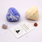 Two gemstone bath bombs, a card with branding, and a small brown object on a light background