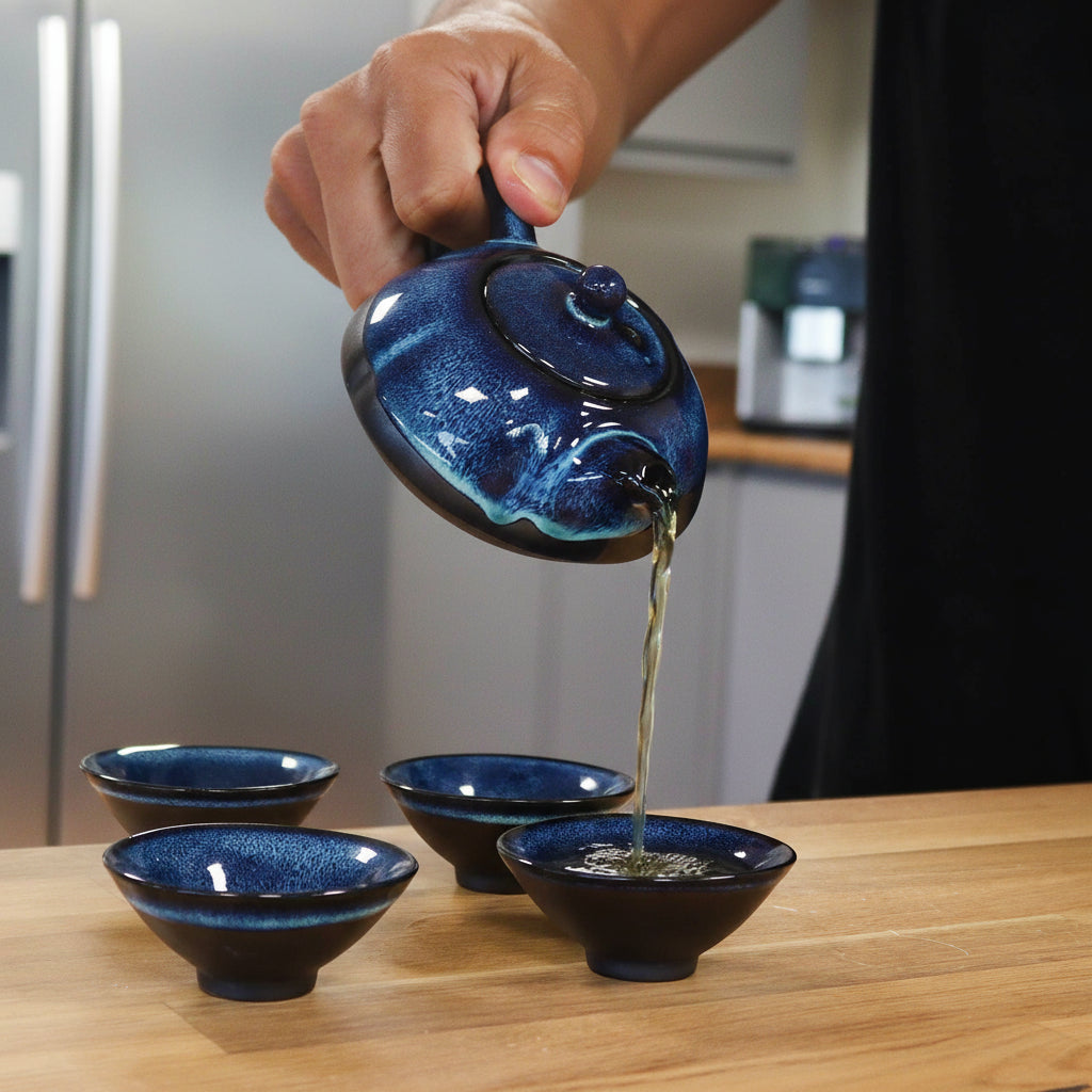 Person pouring tea from a blue ceramic teapot into four matching bowls on a wooden surface.