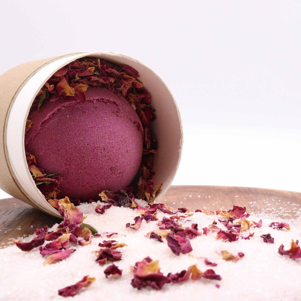 Purple bath bomb in a cardboard tube with rose petals and bath salt on a white background