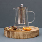 Clear glass teapot with metal mesh on a wooden coaster with dried oranges and flowers against a gray background