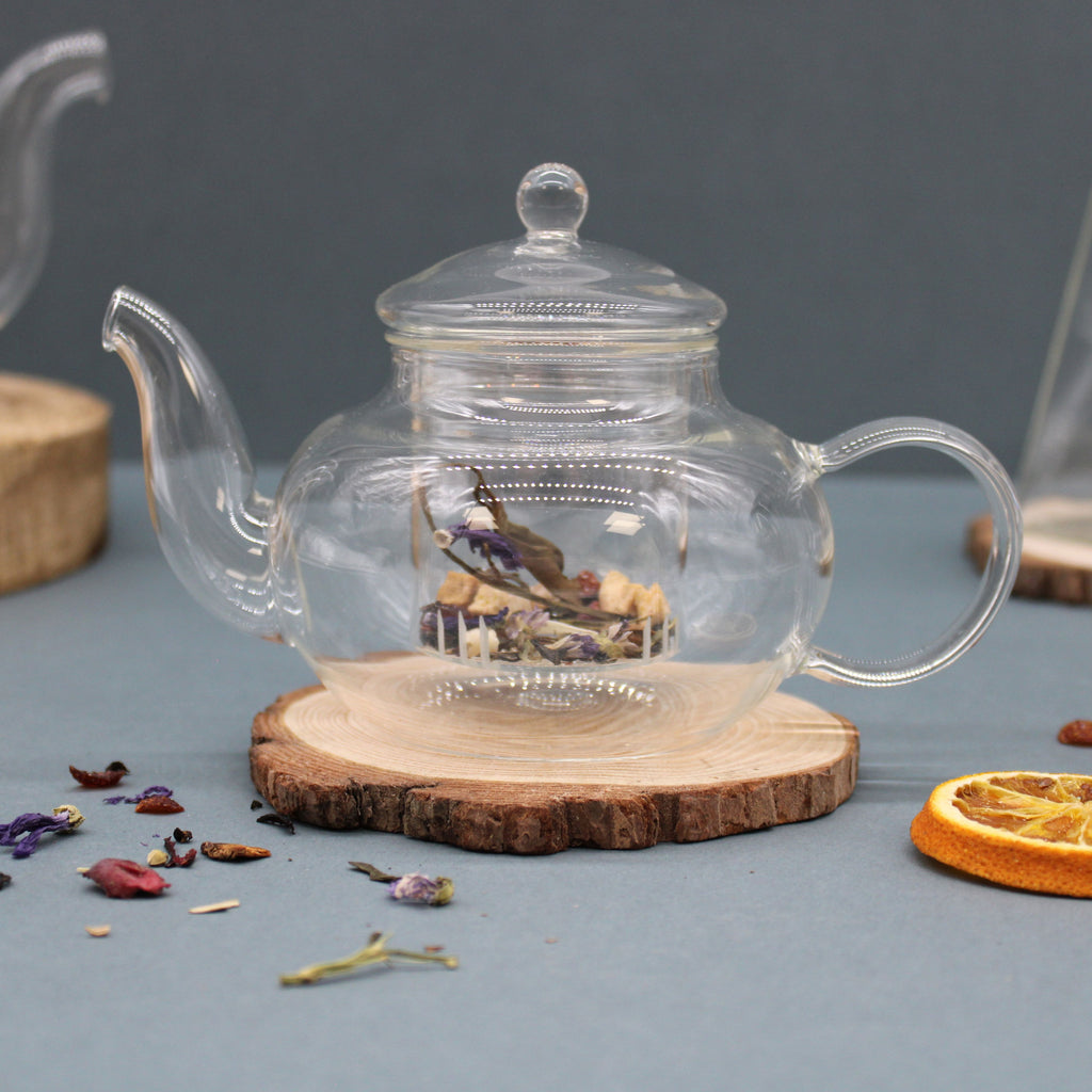 Clear glass teapot with dried herbs on a wooden coaster against a gray background