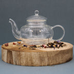 Clear glass teapot on a wooden stand with loose tea leaves on a gray background