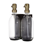Two clear glass bottles with gold pumps in a black holder on a white background