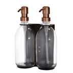 Two clear glass bottles with copper pumps in a black holder on a white background
