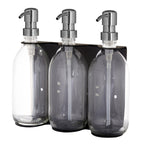Set of three clear glass bottles with silver pumps in a black holder on a white background