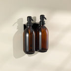 Two amber glass bottles with black pumps on a beige wall-mounted holder against a beige background.