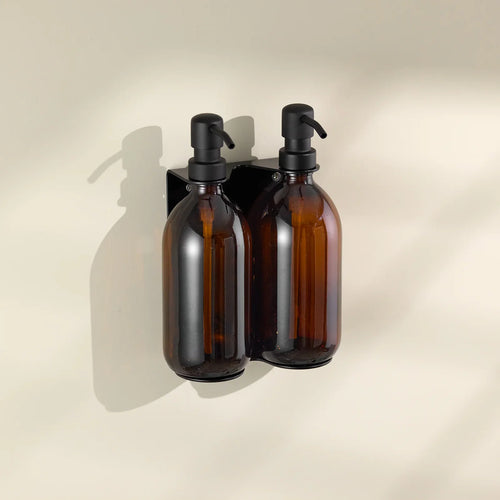 Two amber glass bottles with black pumps on a beige wall-mounted holder against a beige background.