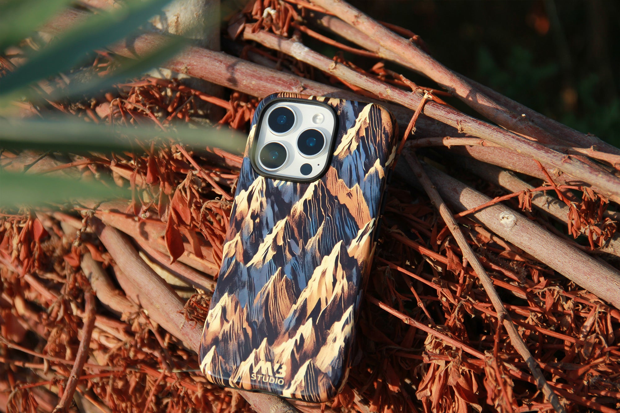 A phone case with a mountain-patterned design in blue, beige, and brown, shown on a smartphone resting on a bed of dry brown branches outdoors. Photograph by MVB STUDIO official store.
