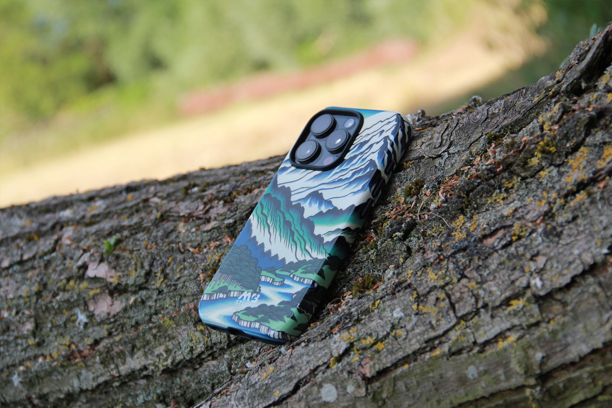 A phone case with a detailed landscape print of mountains, forests, and water, shown resting on a textured tree trunk outdoors. Photograph by MVB STUDIO official store.