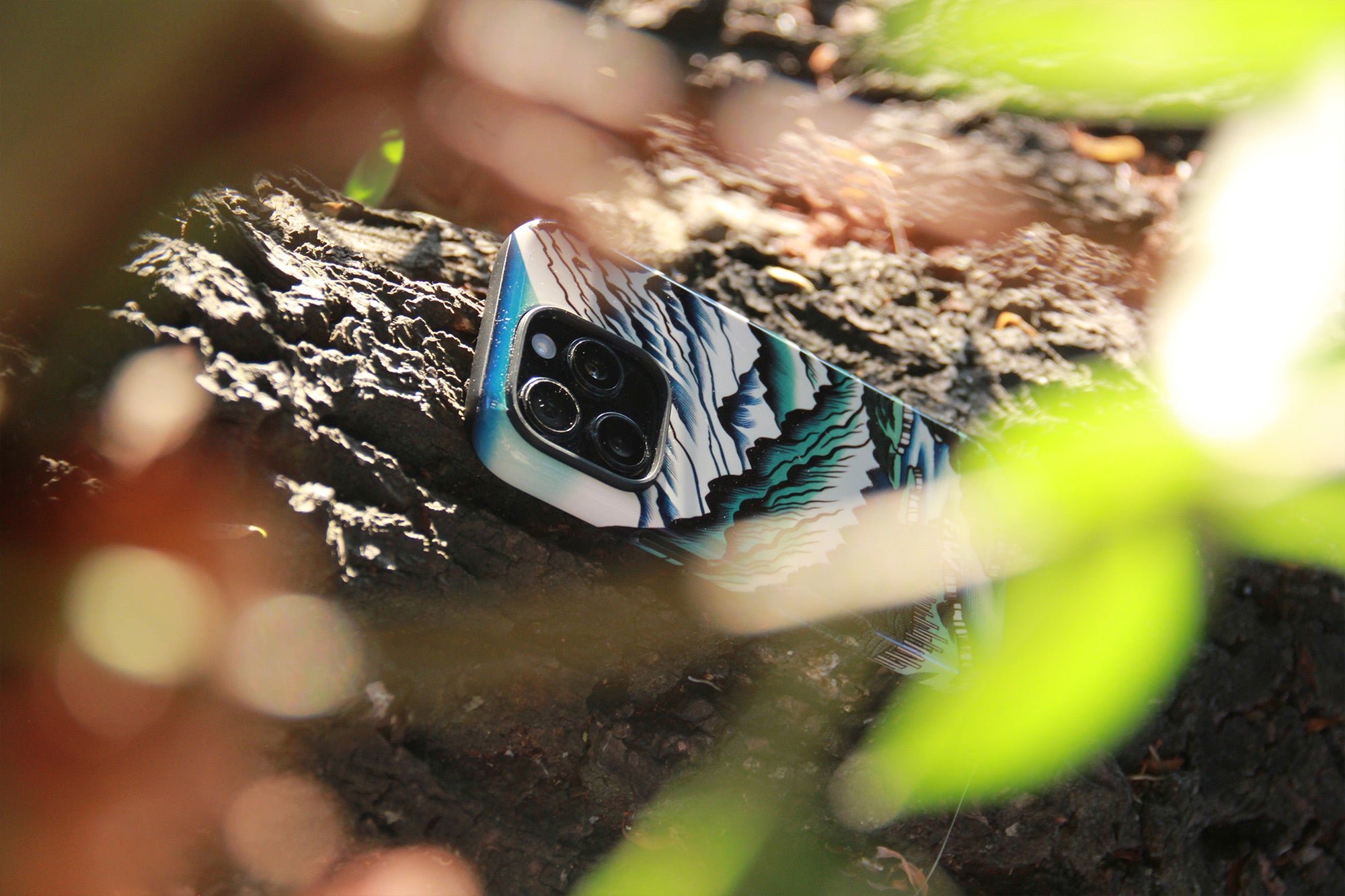 A smartphone case with a blue, white, and green mountain landscape pattern, shown on a phone nestled in rough tree bark outdoors. Photograph by MVB STUDIO official store.