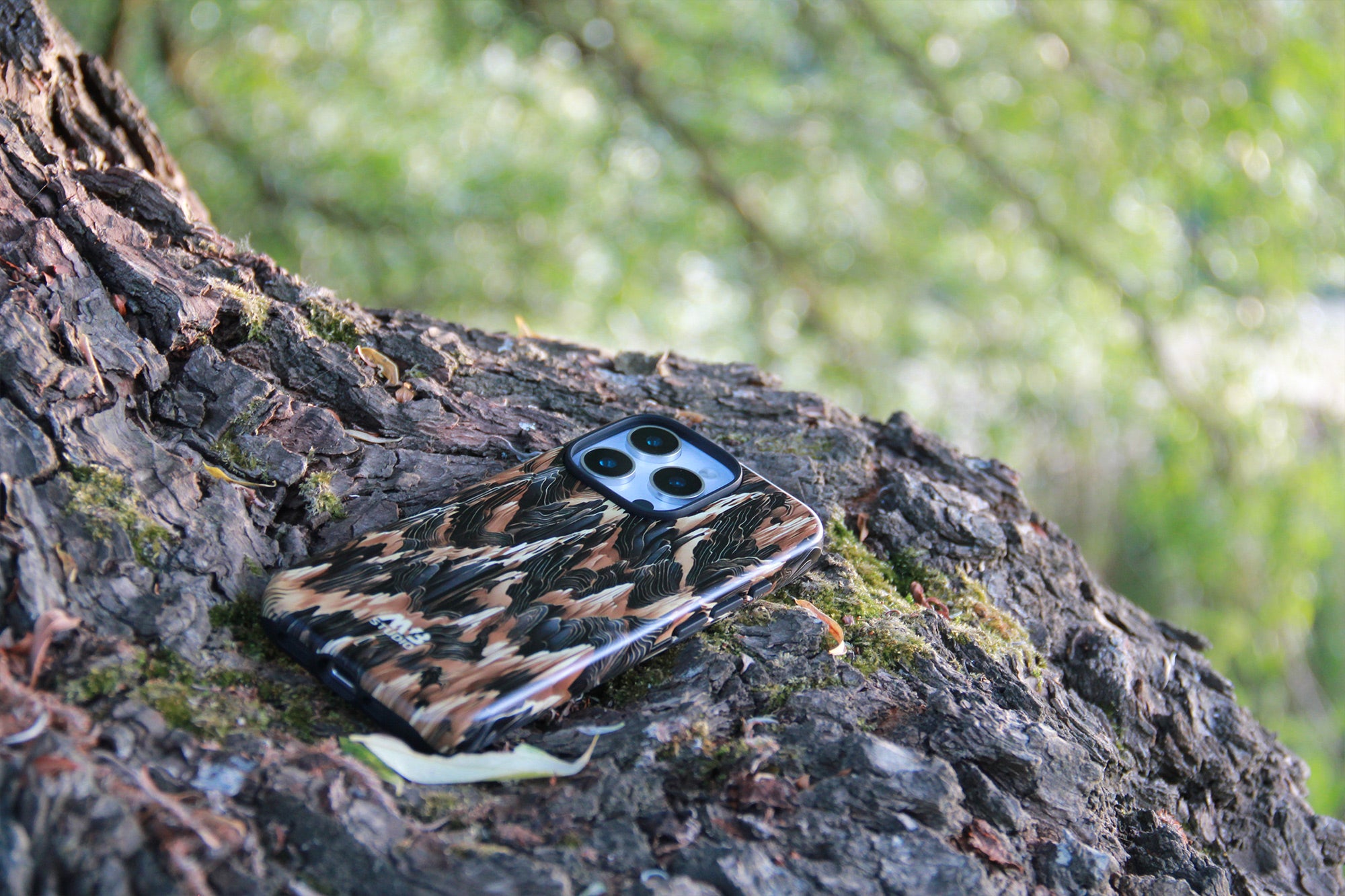 A smartphone case with a brown, black, and beige tiger-stripe camouflage pattern, shown on a tree trunk in an outdoor forest setting. Photograph by MVB STUDIO official store.
