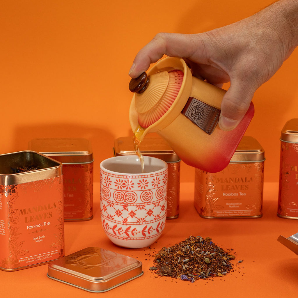 Hand pouring tea from a yellow teapot into a white mug with red patterns on an orange background.