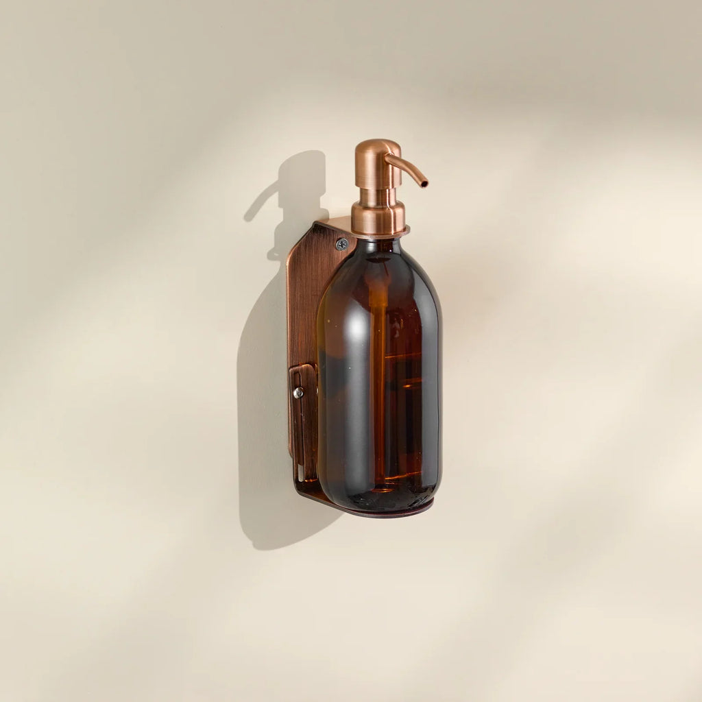 Brown glass soap dispenser with copper pump on a beige wall.