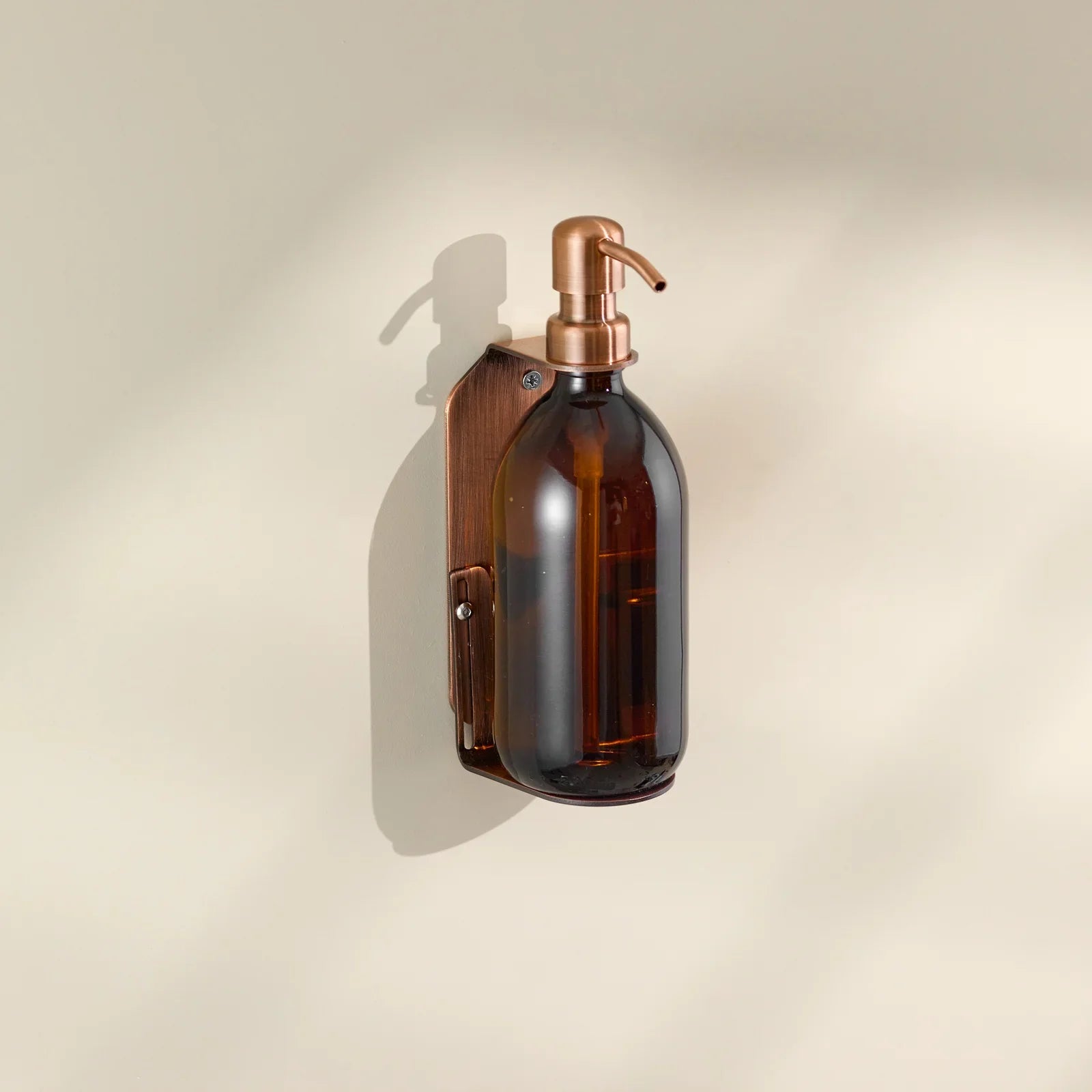 Brown glass soap dispenser with copper pump on a beige wall.