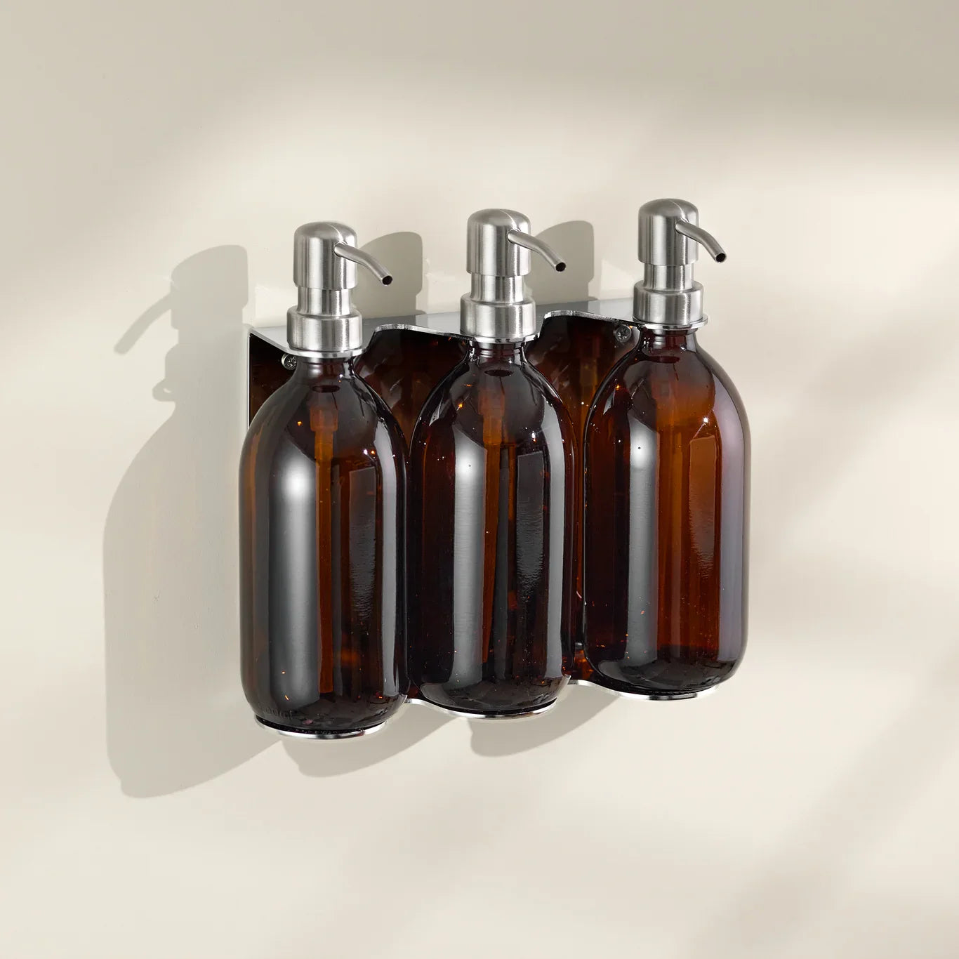 Three amber glass bottles with silver pumps mounted on a beige wall.